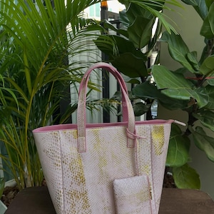 May include: A light pink tote bag and matching wallet with a snakeskin pattern and gold accents. The tote has a rectangular shape with a pink interior and handles. The wallet has a zip closure.