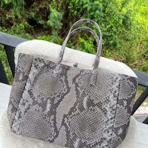 Könnte beinhalten: Graue Tragetasche mit Schlangenmuster. Die Tasche hat zwei Griffe und eine rechteckige Form. Sie liegt auf einer weißen, flauschigen Oberfläche. Der Hintergrund ist unscharf, mit grünem Laub.