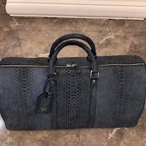 May include: A dark blue duffel bag with a textured snakeskin pattern. The bag has two top handles, a zipper closure, and a small tag with the initials "SN". The bag is approximately 61 cm long.