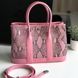 Może przedstawiać: Różowa torebka z wzorem skóry węża i pasującymi różowymi skórzanymi uchwytami i wykończeniami. Torba zawiera odpinany różowy pasek na ramię. Torba stoi na białej powierzchni.