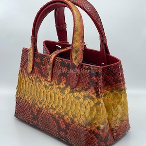 May include: A snakeskin-patterned handbag in red, yellow, and orange. The bag has a structured, rectangular shape with a top handle and contrasting red handle and trim. The bag's exterior showcases a detailed, textured pattern.