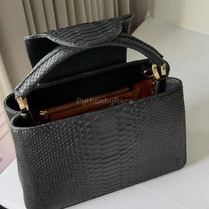 May include: Black handbag with a snakeskin texture. The bag features a top handle and a flap closure. The interior is visible, showing a brown lining and a small zipped pocket. The bag is open, displaying its spacious design.