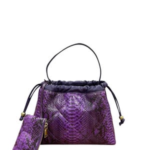 May include: A purple snakeskin-patterned handbag with a matching wallet. The handbag has a drawstring closure and a black handle. The wallet has a gold zipper. The bag and wallet are a deep purple color.