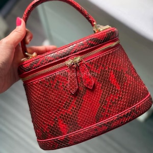 Może przedstawiać: Czerwono-brązowa kosmetyczka z wzorem skóry węża, z górnym uchwytem i złotymi zamkami błyskawicznymi. Cylindryczna torba ma strukturalny kształt i pasujący czerwony suwak. Torba jest trzymana przez osobę.