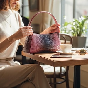 May include: A pink and blue snakeskin-effect handbag with a curved handle. The bag is held by a person at a wooden table. A teacup and saucer, sunglasses, and a smartphone are also on the table.