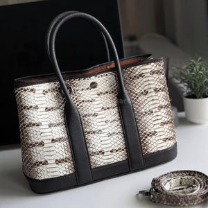 Könnte beinhalten: Eine Handtasche mit Schlangenmuster und schwarzen Lederakzenten. Die Tasche hat ein strukturiertes Design mit einem oberen Griff und einem abnehmbaren Schultergurt. Das Äußere zeigt ein Muster aus weißen, braunen und beigefarbenen Schuppen.
