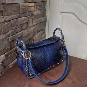 May include: A blue handbag with a snakeskin pattern. The bag has a top handle and a shoulder strap. Gold-coloured rings connect the handles and strap to the bag. The bag is on a wooden surface.