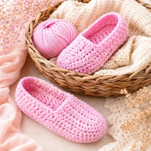 Puede incluir: Un par de zapatillas de ganchillo de color rosa claro con un patrón de puntos texturizado. Una zapatilla está colocada dentro de una cesta redonda tejida junto a un ovillo de lana rosa, mientras la otra descansa frente a la cesta.