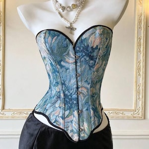 May include: A blue floral patterned corset with a sweetheart neckline and black trim. The corset has a button-up front and is displayed on a mannequin. A pearl necklace with a cross pendant is also visible.