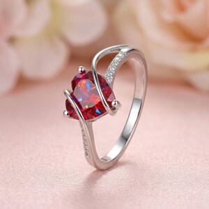 Könnte beinhalten: Ein silberner Ring mit einem herzförmigen, facettierten roten Edelstein. Der Ring hat ein zartes Design mit kleinen, klaren Akzentsteinen und einer polierten Oberfläche. Der Edelstein ist der Mittelpunkt, der in einem modernen, eleganten Stil gefasst ist.