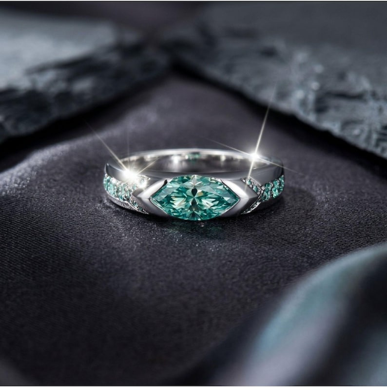 May include: A silver ring with a large, marquise-cut, teal-colored gemstone as the centerpiece. Smaller teal stones are set along the sides. The ring is set against a dark, textured background, with light reflecting off the ring's surface.