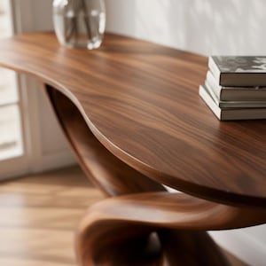 May include: A contemporary console table crafted from dark wood, showcasing a distinctive, fluid form. The table has a curved top and base, with a natural wood grain. A stack of books and a clear vase are on the table's surface.