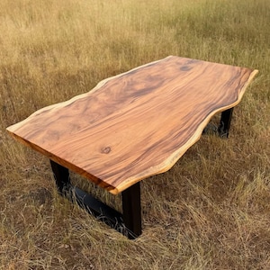 Op de afbeelding: Een rechthoekige houten tafel met een levende rand en een natuurlijke houtnerf afwerking. De tafel wordt ondersteund door zwarte metalen poten. De tafel staat buiten in een veld met hoog gras. De tafel is ongeveer 180 cm lang.