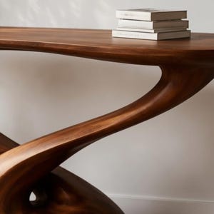 May include: A dark brown wooden console table with a distinctive, flowing design. The table has a smooth, polished finish and a unique, organic shape. Three books with white covers are stacked on the table's surface. The table is a piece of modern furniture.