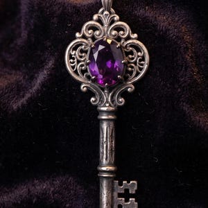 Op de afbeelding: Een zilverkleurige metalen hanger in de vorm van een oude sleutel, voorzien van een ovaal geslepen dieppaarse edelsteen in de versierde kop. De schacht van de sleutel heeft een getextureerde, verweerde afwerking en de baard is rechthoekig.