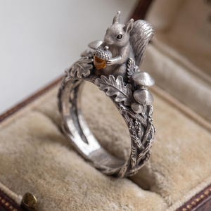 May include: A silver ring featuring a sculpted squirrel holding a small amber-colored acorn. The band is detailed with raised oak leaves, small mushrooms, and textured branches that wrap around the finger in a continuous, intricate forest design.