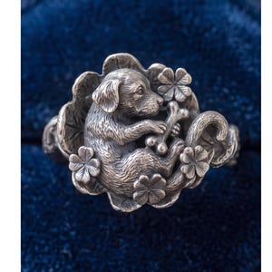 May include: A silver ring featuring a detailed sculptural design of a puppy holding a bone, surrounded by embossed four-leaf clover motifs. The metal has an oxidized finish that highlights the texture of the fur and the intricate floral patterns.