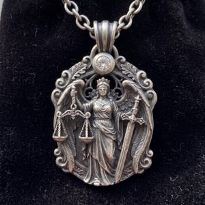 May include: A silver pendant necklace featuring a detailed relief of a blindfolded winged figure wearing a crown, holding a set of scales in one hand and a sword in the other. The oval-shaped pendant has an ornate border and a single clear stone.