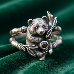 May include: A silver ring featuring a detailed panda bear design. The panda is holding bamboo, and a black gemstone is set in the center. The ring has a textured finish and is set against a green background.