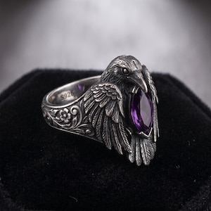 May include: A sterling silver ring featuring a detailed raven with spread wings and a marquise-cut purple gemstone held in its beak. The band is decorated with floral engravings and is stamped with S925 on the inner surface of the metal.