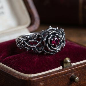 May include: A silver ring featuring a detailed rose design with thorny vines wrapping around the band. The center of the rose is set with a small, round, deep red gemstone. The metal has an oxidized finish to highlight the intricate textures.