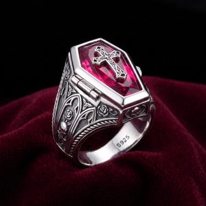 Puede incluir: Un anillo de plata con forma de ataúd y tapa abatible, con una piedra roja facetada incrustada y una cruz de plata encima. Los laterales presentan relieves de calaveras y rosas, y el interior de la banda lleva el sello S925.