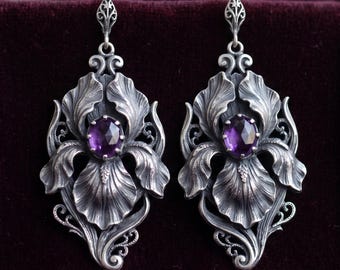 May include: A pair of sterling silver drop earrings featuring an intricate, three-dimensional iris flower design. Each earring is centered with a faceted, oval-shaped purple amethyst gemstone and hangs from a decorative, filigree-style hook.