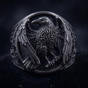 May include: A detailed, silver-toned ring featuring a raven with outstretched wings. The bird is intricately carved with ornate feather and wing details. The ring has a dark, textured background, highlighting the detailed craftsmanship of the jewelry.