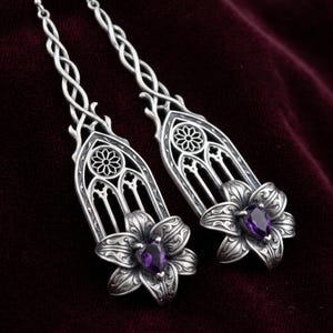 Può includere: Un paio di lunghi orecchini in argento con un intricato design ad arco gotico e un motivo a rosone circolare sulla parte superiore. Ogni orecchino termina con un fiore di giglio in argento scolpito con un'ametista viola a forma di goccia.