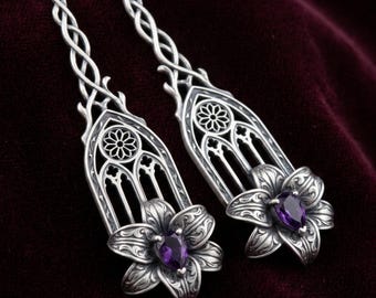May include: A pair of long silver earrings featuring an intricate gothic arch design with a circular rose window pattern at the top. Each earring terminates in a sculpted silver lily flower with a teardrop-shaped purple amethyst gemstone at the center.