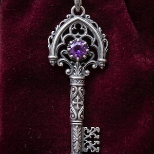 Op de afbeelding: Een zilveren hanger in de vorm van een versierde sleutel met een ronde, gefacetteerde paarse edelsteen in de kop. Het metaalwerk bevat ingewikkeld filigraan, kruismotieven en bloemmotieven langs de schacht en een gedetailleerde baard.