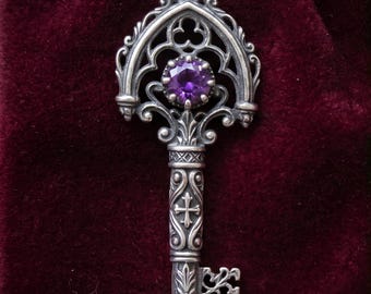 May include: A silver pendant shaped like an ornate key, featuring a round, faceted purple gemstone set in the bow. The shank is decorated with intricate relief carvings, including a cross and floral motifs, with a stamped 925 mark on the bail.