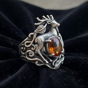 May include: A silver ring featuring a detailed deer design with an oval, amber-colored gemstone. The ring has an ornate, nature-inspired pattern around the band and the deer is sculpted with intricate detail. The gemstone is approximately 0.75 inches long.