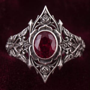 May include: A silver ring featuring an oval-cut deep red gemstone set in the center. The band and setting are crafted with intricate metalwork depicting sculpted rose blossoms and detailed leaves, creating a pointed, symmetrical floral frame.
