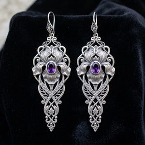 May include: A pair of long silver drop earrings featuring an intricate floral design with swirling metal filigree patterns. Each earring is centered with a single oval-cut purple amethyst gemstone and hangs from a decorative silver hook wire.