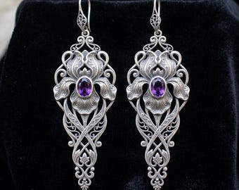 May include: A pair of long silver drop earrings featuring an intricate floral design with swirling metal filigree patterns. Each earring is centered with a single oval-cut purple amethyst gemstone and hangs from a decorative silver hook wire.