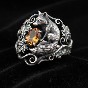 May include: A sterling silver ring featuring a sculpted fox figure perched beside an oval-cut amber-colored gemstone. The band is decorated with intricate metalwork depicting detailed maple leaves and swirling vine patterns in a dark oxidized finish.