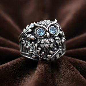 May include: A silver ring featuring a detailed owl design with two round, light blue gemstone eyes. The band is decorated with sculpted oak leaves and small acorns, finished with an oxidised texture to highlight the intricate metalwork details.