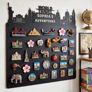 Op de afbeelding: Een zwart metalen displaybord met de tekst "WORLD TRAVEL SOPHIA'S ADVENTURES" en silhouetten van steden. Het bord bevat diverse souvenirmagneten, waaronder bezienswaardigheden zoals de Golden Gate Bridge en de Eiffeltoren.
