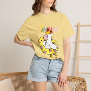 Może przedstawiać: Jasnożółty t-shirt z krótkim rękawem z grafiką przedstawiającą białą kaczkę w okularach przeciwsłonecznych w kształcie serc, siedzącą w żółtym kole do pływania w kwiaty. Kaczka trzyma różowego loda. Powyżej znajduje się napis SUMMER kolorowymi literami.