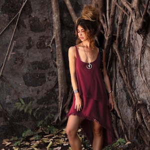 May include: A woman wearing a burgundy sleeveless dress with a fringe hem, standing in front of a tree with exposed roots. She is wearing a silver necklace with a circular pendant.