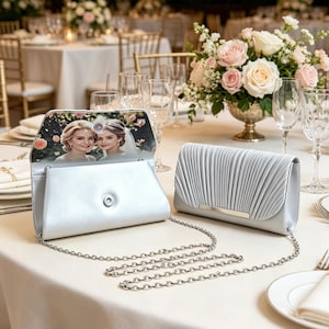 Könnte beinhalten: Zwei silberne Clutch-Taschen mit abnehmbaren Kettenriemen. Eine Tasche ist geöffnet und zeigt ein aufgedrucktes Foto von zwei Personen auf der Innenseite. Die andere Tasche ist geschlossen und hat ein plissiertes Design mit Metallakzent.