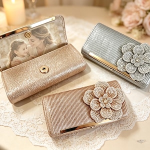 May include: Three rectangular clutch bags with metallic finishes. One rose gold clutch is open, displaying a photograph of a person and a child inside. Two closed clutches, one silver and one rose gold, feature large, sparkling floral embellishments.