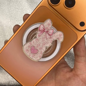 May include: A gold-colored phone case with a bunny-shaped phone grip. The bunny is decorated with a floral pattern and a pink bow and heart. The phone has three camera lenses.