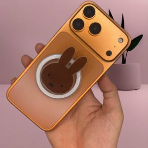 May include: A gold-coloured phone case featuring a brown bunny design. The case has a clear, transparent section with a brown bunny graphic and a white ring. The phone case is held in a hand.