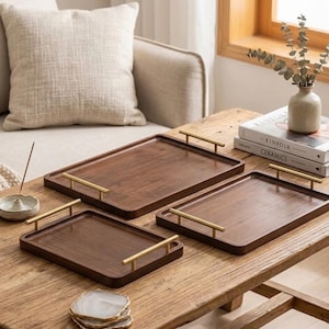 May include: Three rectangular dark brown wooden serving trays of varying sizes, each featuring two horizontal brushed gold metal handles attached to the short sides. The trays have a smooth finish and are arranged on a wooden surface.