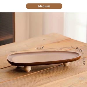 May include: An oval wooden tray with a smooth brown finish, supported by three small, light-colored rounded feet. The tray features dimensions of 11.8 inches in length, 5.0 inches in width, and 2.0 inches in height, labeled as Medium.