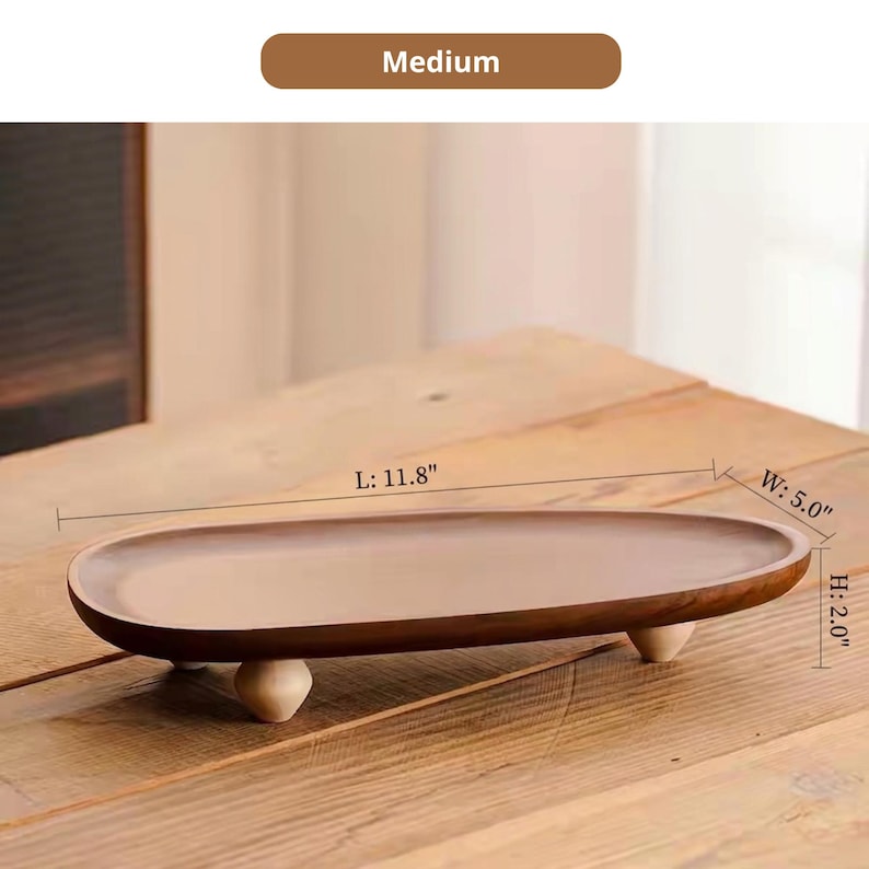 May include: An oval wooden tray with a smooth brown finish, supported by three small, light-colored rounded feet. The tray features dimensions of 11.8 inches in length, 5.0 inches in width, and 2.0 inches in height, labeled as Medium.