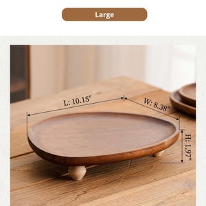 May include: A large, oval-shaped wooden tray with a smooth, dark brown finish, supported by three small, light-colored wooden feet. The tray features printed dimensions of 10.15 inches in length, 8.38 inches in width, and 1.97 inches in height.