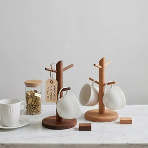 May include: Two wooden mug trees, one dark walnut and one light wood, each holding white ceramic mugs. A glass jar with a wooden lid and brass clips sits to the left. The tag on the dark walnut tree reads "Handcrafted Solid Walnut Portland, OR."